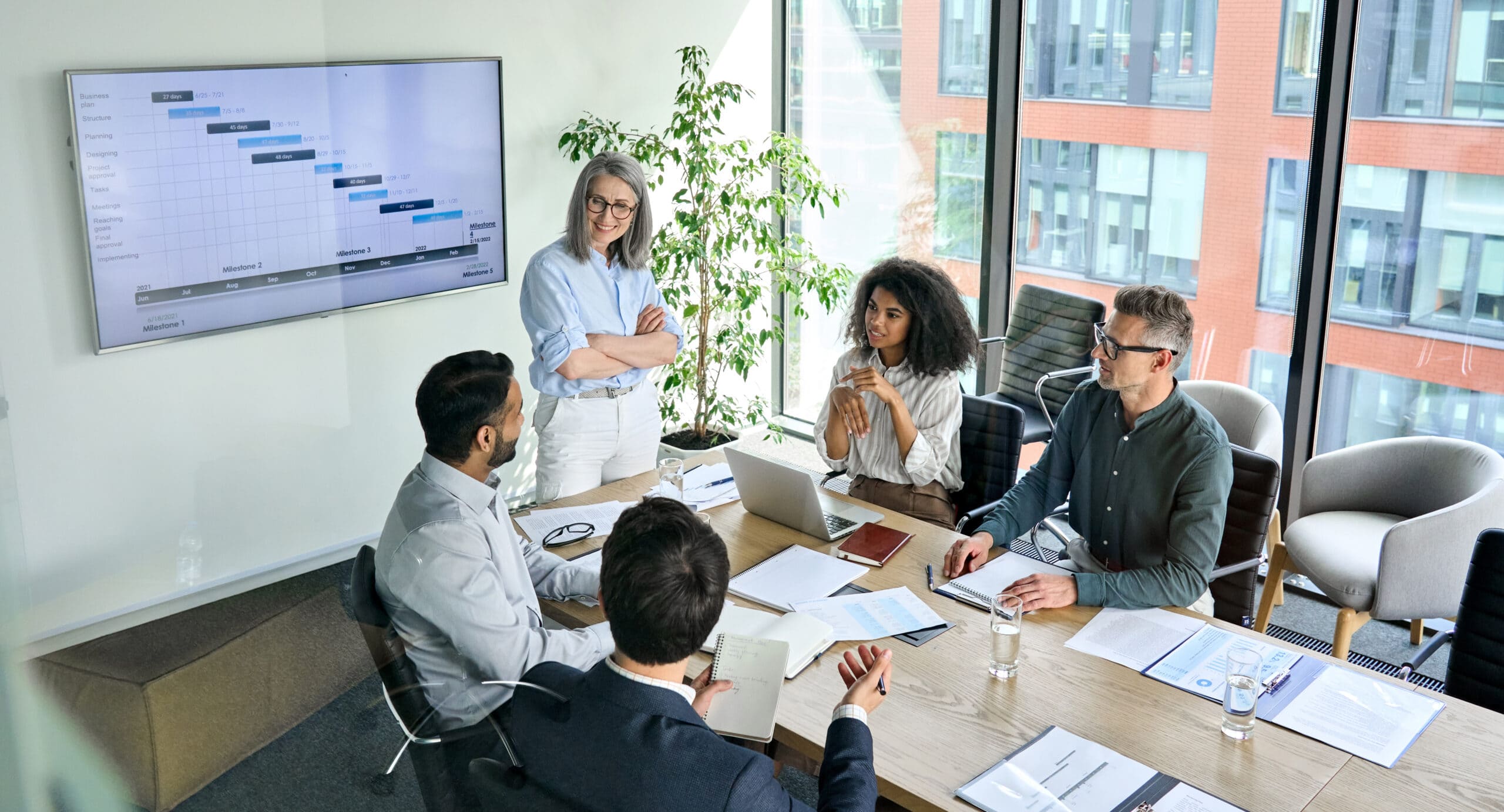 Diverse corporate team working together in modern meeting room office. A professional meeting in a modern office setting. Five people are gathered around a table, engaged in discussion. One person, an older woman with gray hair and glasses, is standing by a large screen displaying a Gantt chart, which shows project milestones and timelines. She appears to be leading the discussion. The other participants, a mix of men and women, are seated around the table, which has laptops, notebooks, documents, and glasses of water. A large window in the background offers a view of a building outside, and there is a green plant next to the window, adding a natural touch to the space.
