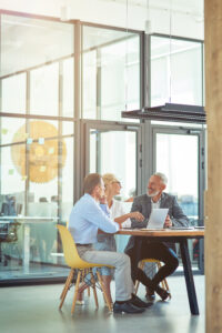 Vertical shot of three mature colleagues sitting in the modern office or coworking space, using laptop and discussing work. Teamwork and cooperation concept