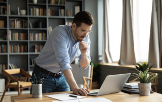 Thoughtful young businessman in eyeglasses working online on computer standing at table, analyzing report, making difficult decisions, developing company growth strategy at modern home office.