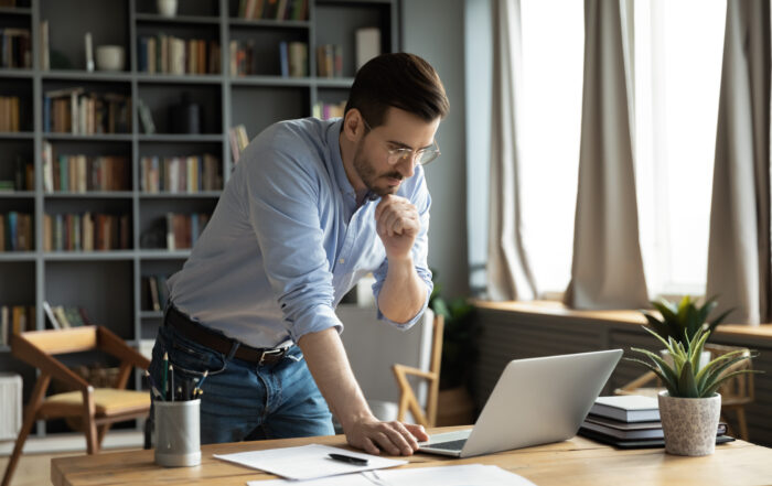 Thoughtful young businessman in eyeglasses working online on computer standing at table, analyzing report, making difficult decisions, developing company growth strategy at modern home office.