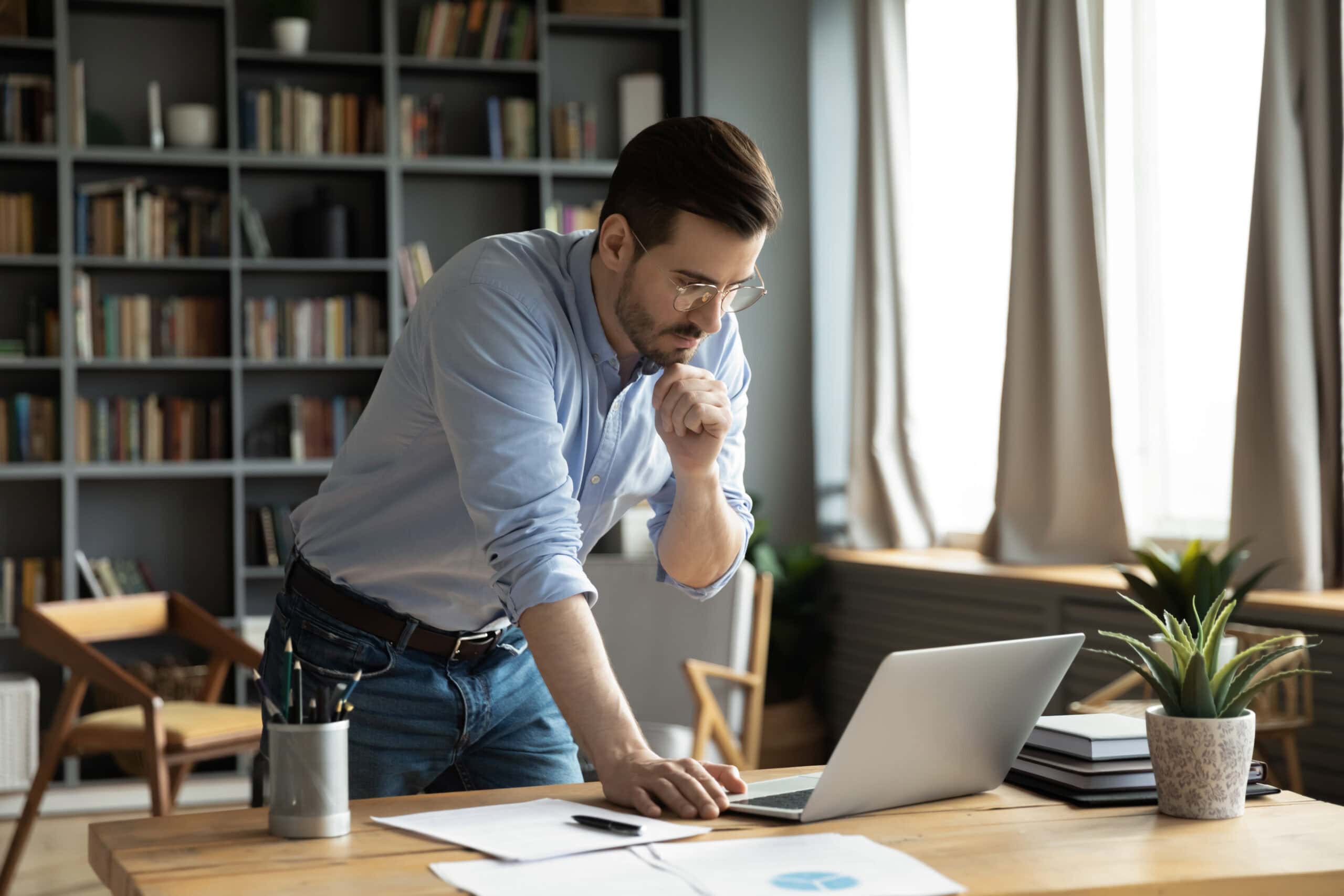 Thoughtful young businessman in eyeglasses working online on computer standing at table, analyzing report, making difficult decisions, developing company growth strategy at modern home office.